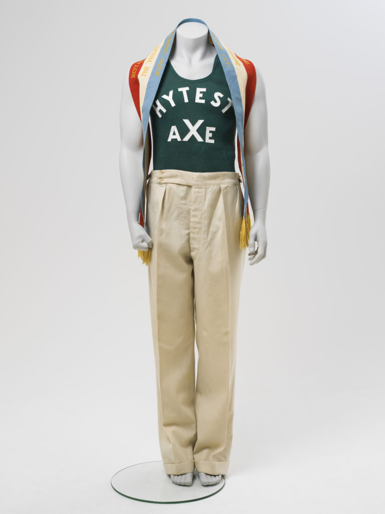 Mannequin wearing a green-coloured cotton ribbed 'Chesty' Bonds Athletic singlet with 'HYTEST AXE' in white vinyl lettering on the front and back, and cream trousers. Around his neck is a championship ribbon for wood chopping. 